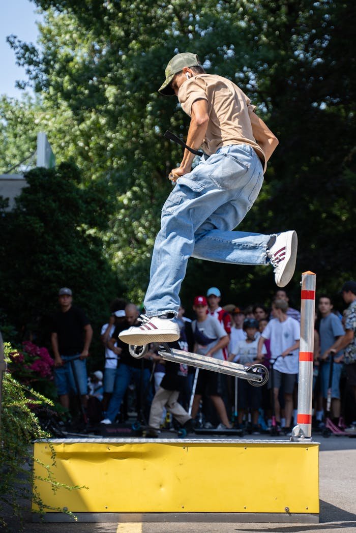 A young man performs an impressive freestyle scooter trick in front of a crowd outdoors.